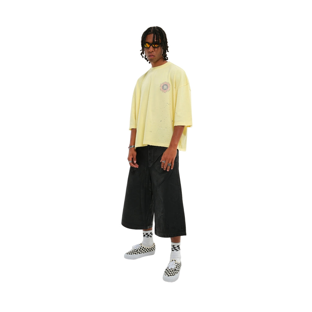 A model wearing a pale yellow oversized t-shirt with a small circular graphic on the left chest, paired with black wide-leg cropped pants, white socks, and black-and-white checkered sneakers, set against a plain white background.