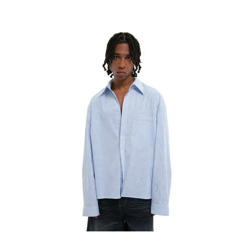 A model with dreadlocks wearing a light blue and white vertically striped button-down shirt, styled open at the collar, paired with dark jeans against a plain white background.