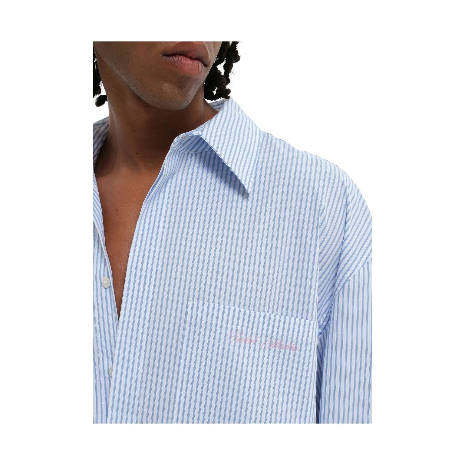 A close-up of a person wearing a light blue and white vertically striped button-down shirt with a chest pocket and subtle pink embroidered text.