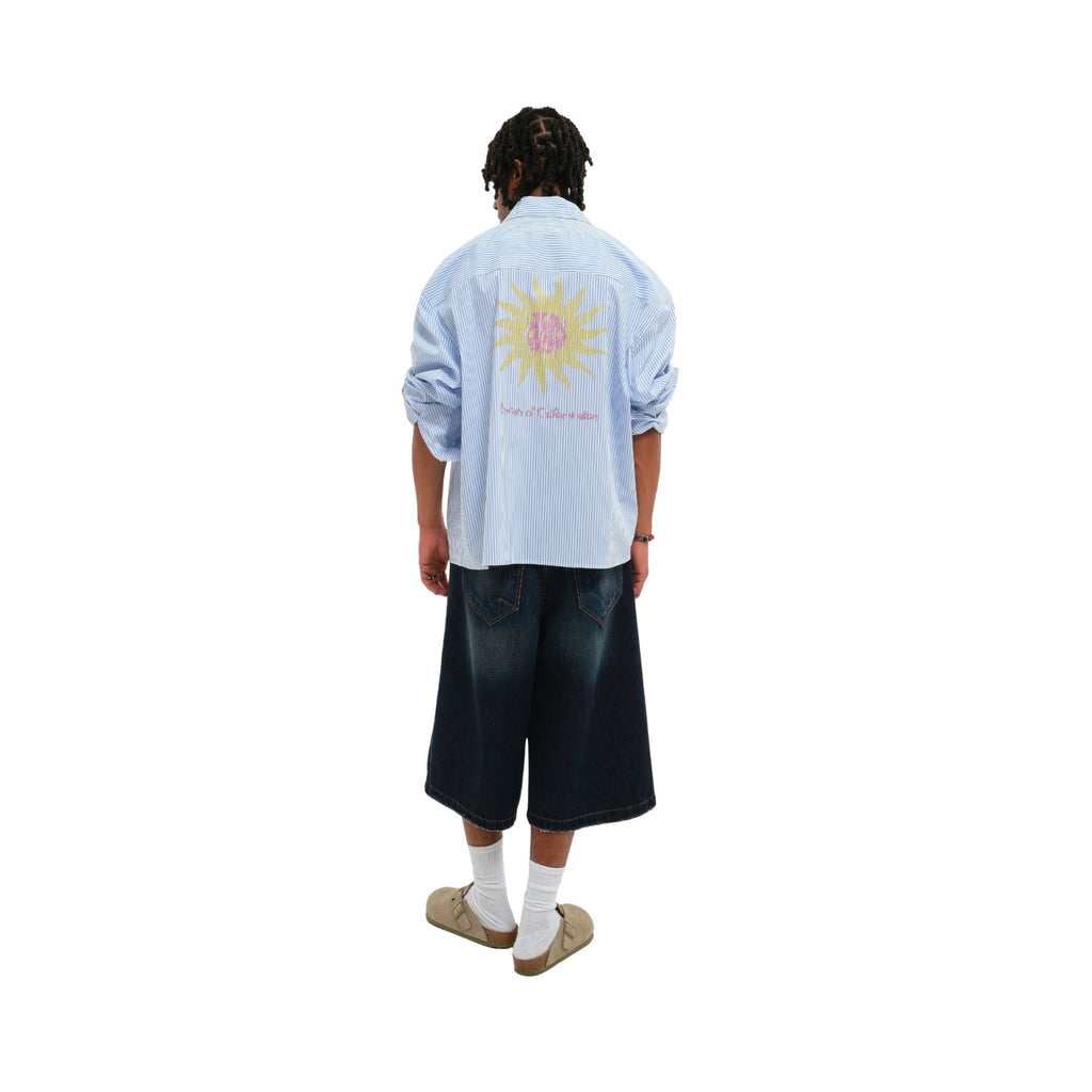 A model shown from the back wearing a light blue striped button-up shirt with a large yellow sun graphic and pink text on the back, paired with dark denim shorts, white socks, and beige shoes, against a plain white background.