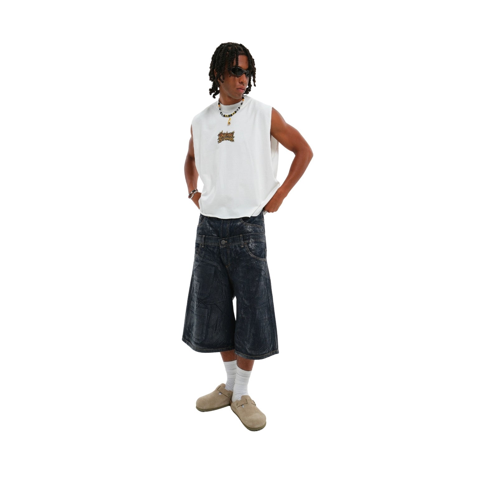 A model wearing a white sleeveless crop top with a small brown graphic on the chest, dark denim shorts, white socks, and beige slip-on shoes, set against a plain white background.
