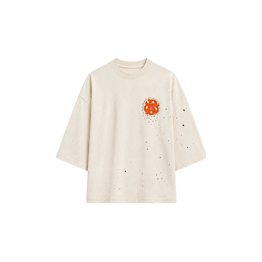 An off-white short-sleeve t-shirt with a small orange graphic on the left chest and scattered black speckles, shown against a plain white background.