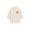 An off-white short-sleeve t-shirt with a small orange graphic on the left chest and scattered black speckles, shown against a plain white background.