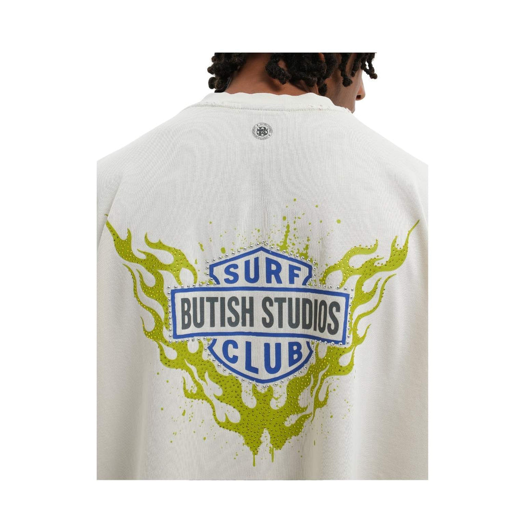 A person shown from the back wearing a white ribbed sweatshirt with a large graphic on the back. The graphic features a blue and white shield reading 'SURF BUTISH STUDIOS CLUB' with lime green flame-like splatter details.