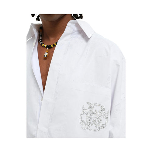 A close-up of a person wearing a white button-up shirt with a silver rhinestone monogram logo on the left chest pocket. The person is also wearing a black and yellow beaded necklace with a small pendant, set against a plain white background.