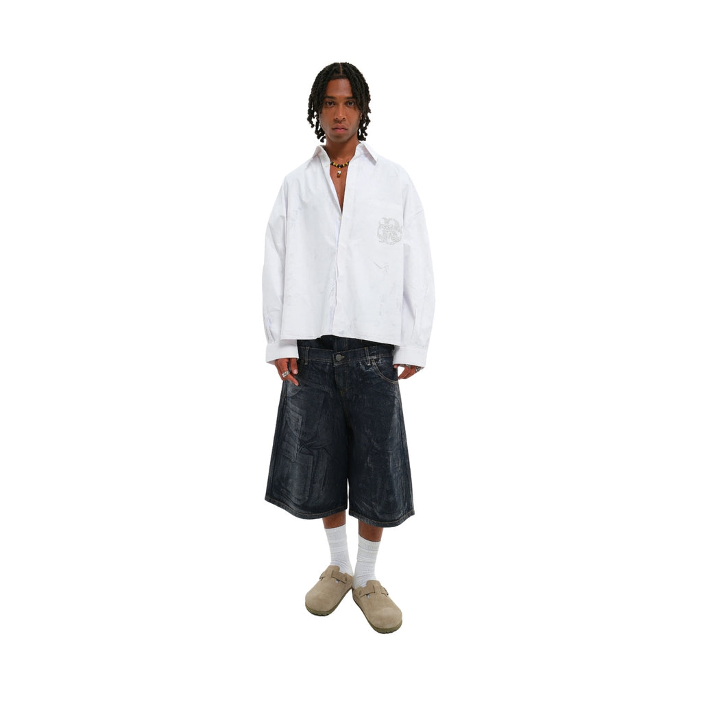 A model stands against a plain white background wearing a white long-sleeve button-up shirt with a tonal chest graphic, dark denim cropped shorts, white socks, and beige slip-on shoes.