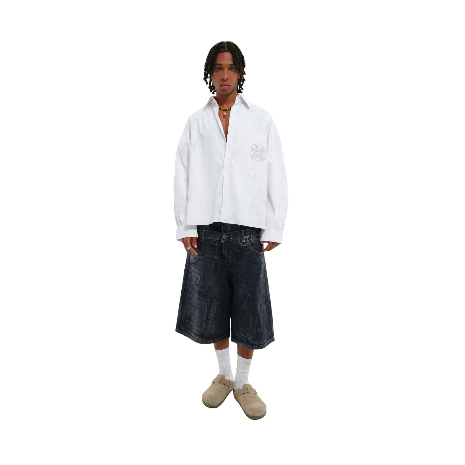 A model stands against a plain white background wearing a white long-sleeve button-up shirt with a tonal chest graphic, dark denim cropped shorts, white socks, and beige slip-on shoes.