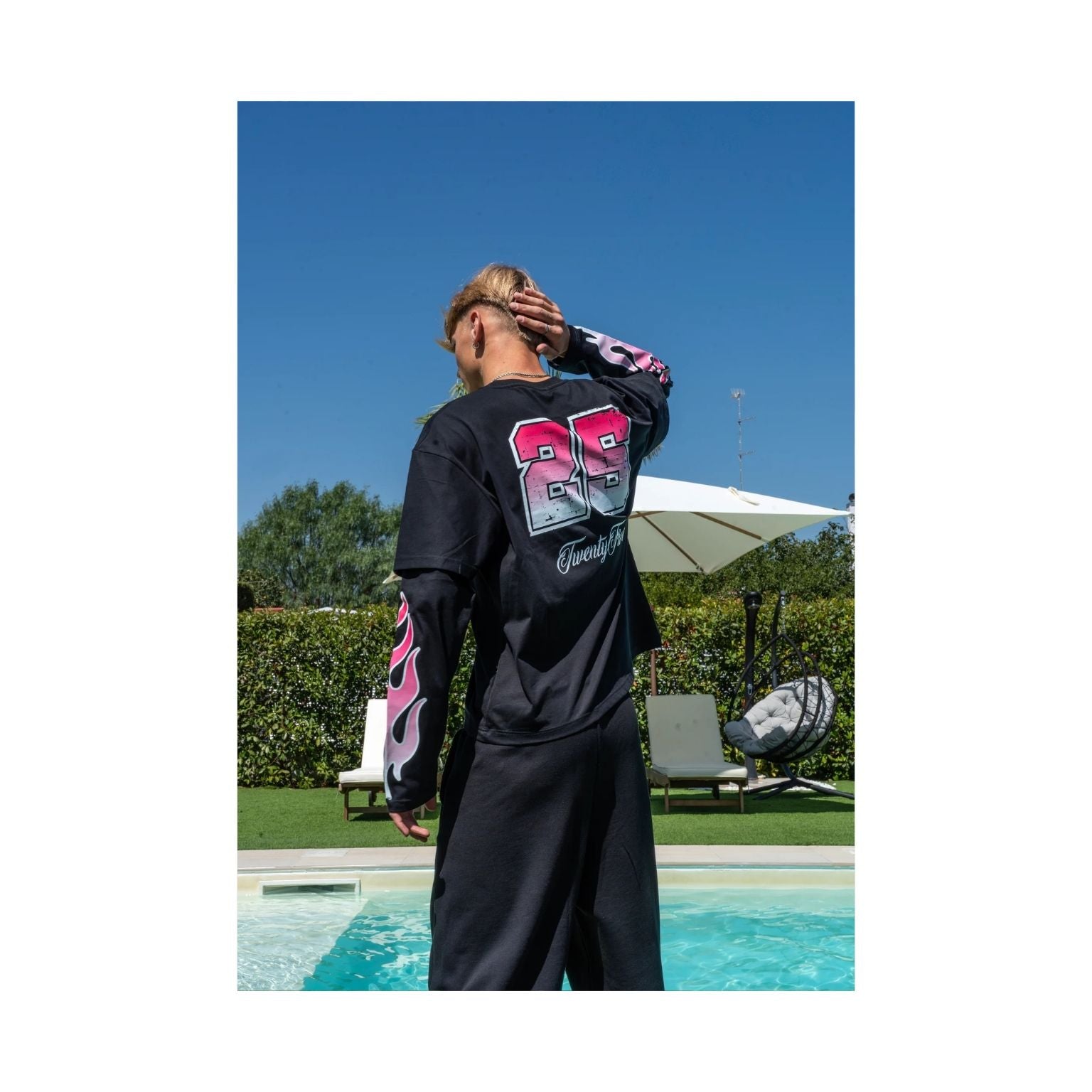 A man stands by a swimming pool outdoors, wearing a black long-sleeve graphic shirt and matching black pants. The shirt features a large red, pink, and gray graphic on the back and pink flame-style graphics on the sleeves.