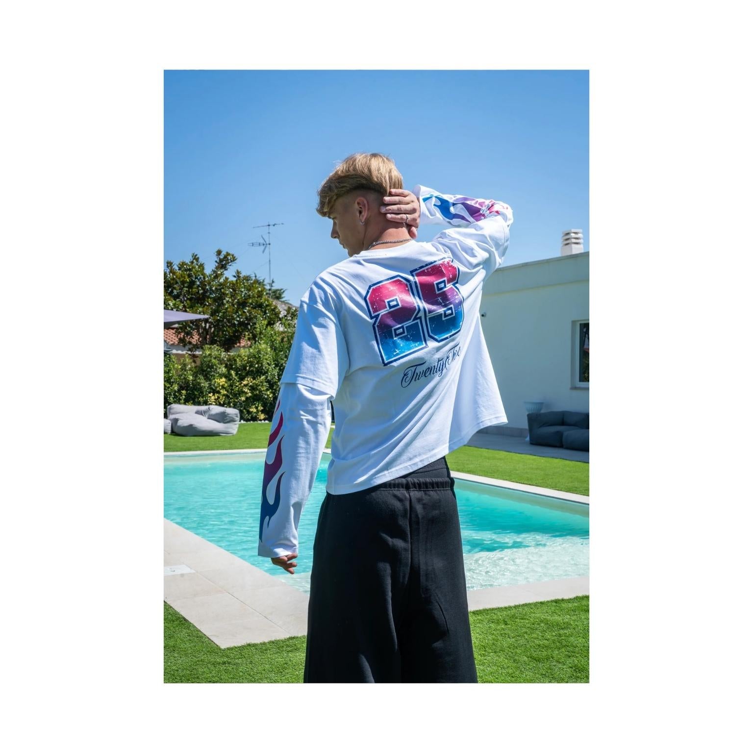 A man stands outdoors by a swimming pool wearing a white long-sleeve t-shirt with a large red, blue, and purple graphic on the back and black pants.