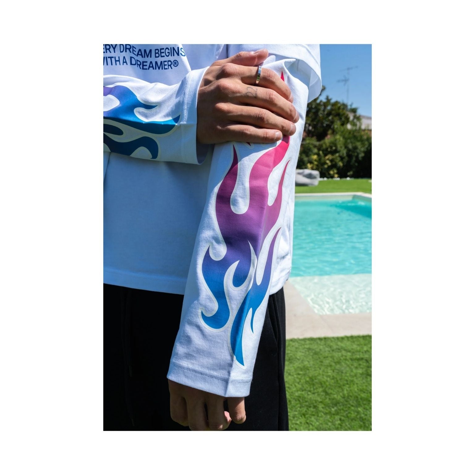 A person outdoors by a swimming pool wearing a white long-sleeve shirt with a large blue, purple, and pink flame graphic printed down one sleeve.