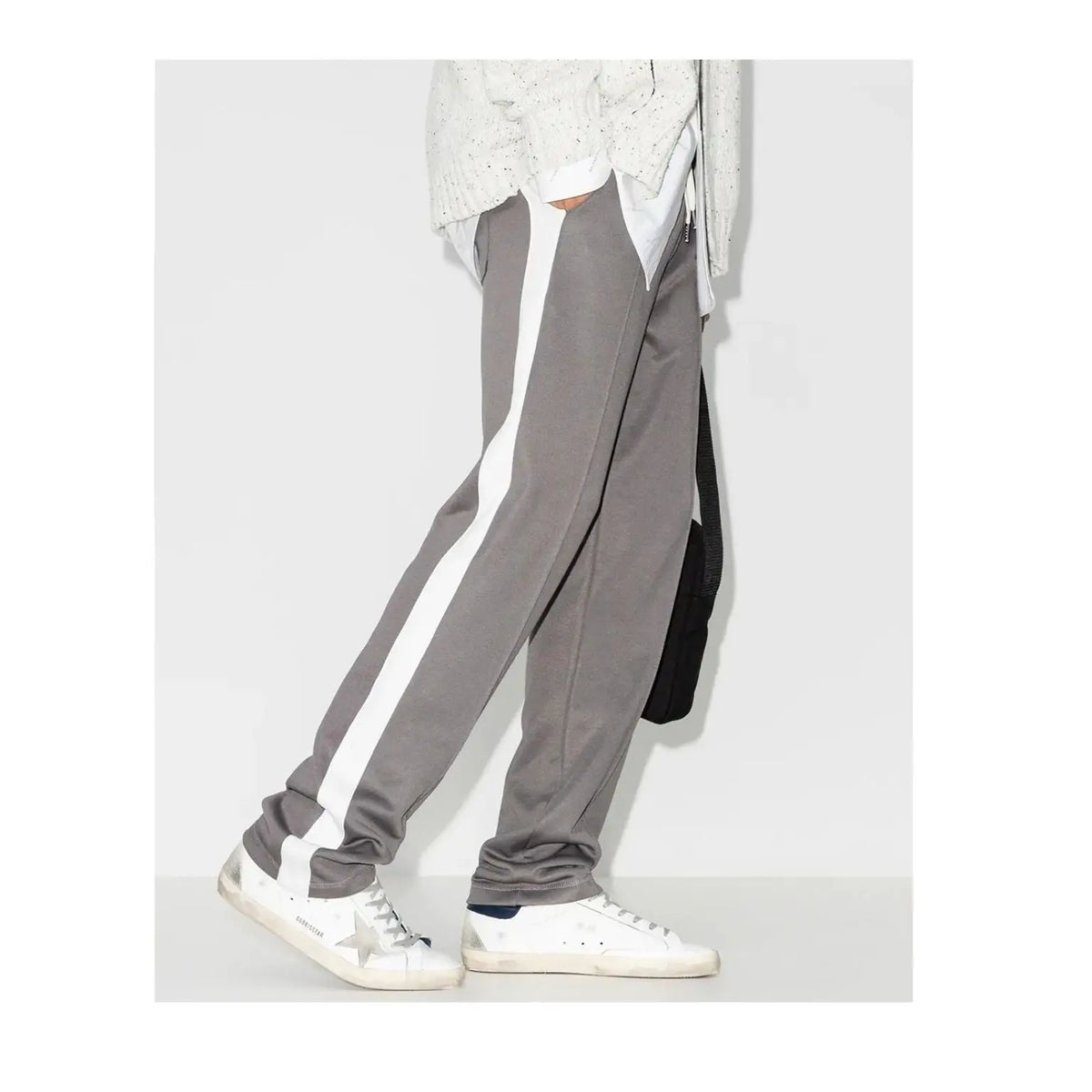 A person wearing gray jogger-style pants with a wide white side stripe, paired with white sneakers and a light knit top, shown from the waist down against a plain light gray studio background.