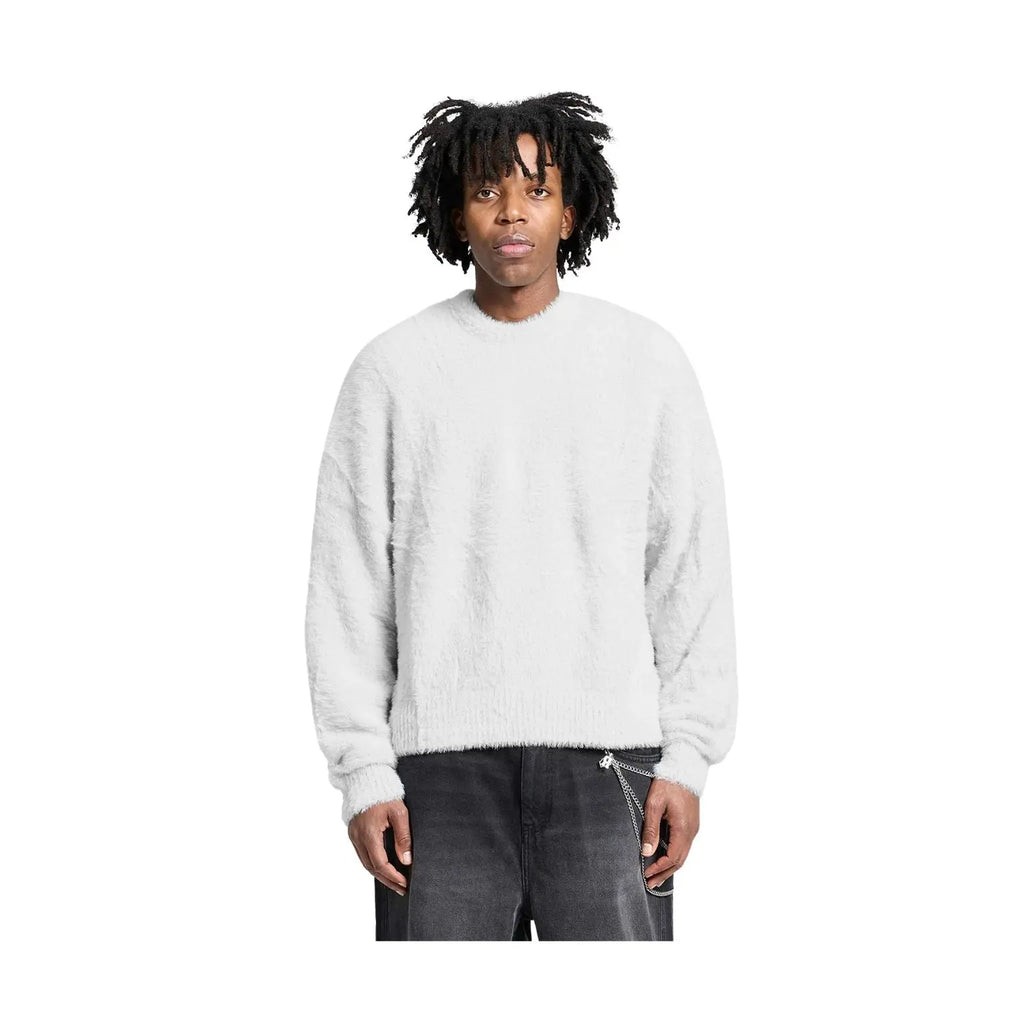 A man with black dreadlocks wearing a light gray fuzzy crewneck sweater and dark gray jeans, standing against a solid white background.