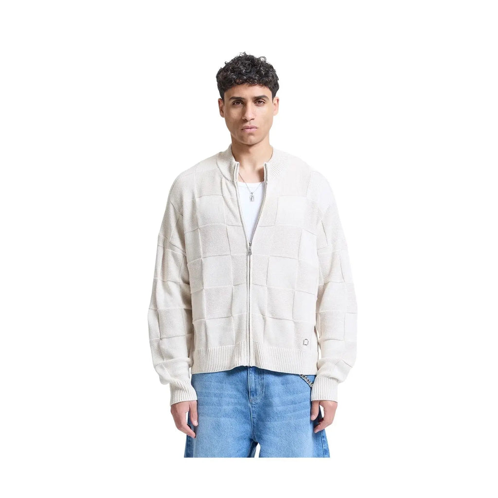 A man wearing a cream-colored zip-up knit cardigan with a patchwork grid pattern, styled with blue jeans and a white t-shirt, shot against a solid white background.