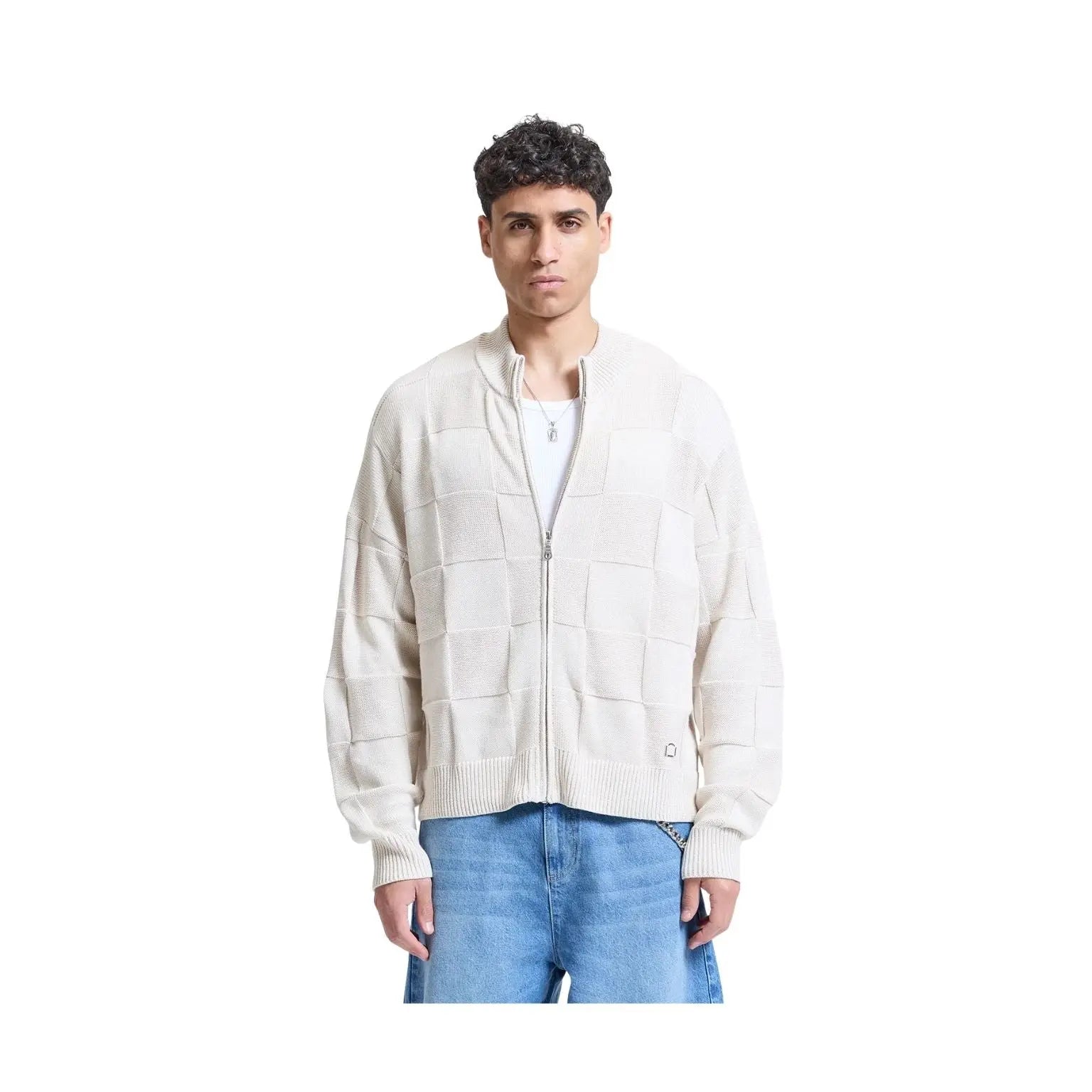 A man wearing a cream-colored zip-up knit cardigan with a patchwork grid pattern, styled with blue jeans and a white t-shirt, shot against a solid white background.